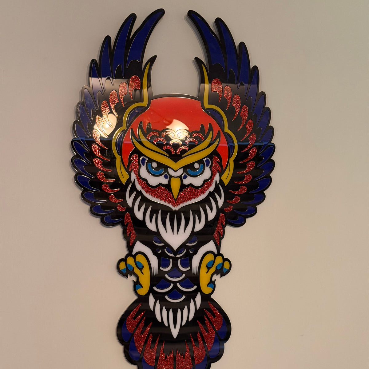 A decorative owl wall art piece with spread wings, featuring a red, blue, white, and yellow design mounted on a plain wall.