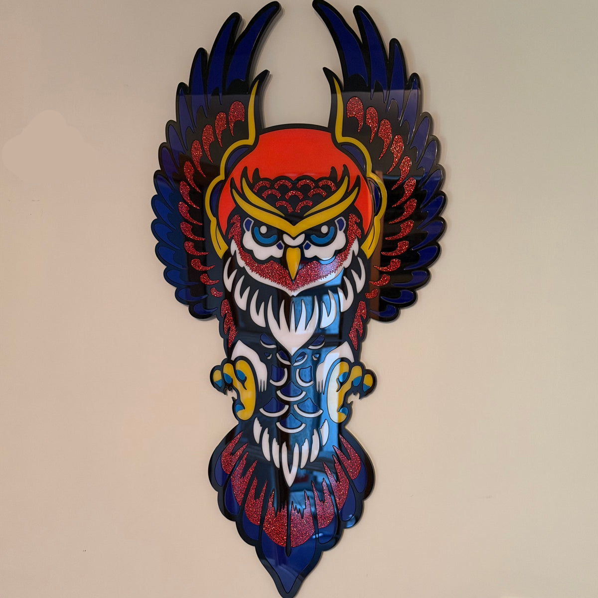 A colorful owl wall art piece mounted on a beige wall, featuring a stylized owl with dark blue wings, a red and yellow head, white feathers, and blue and red body details.