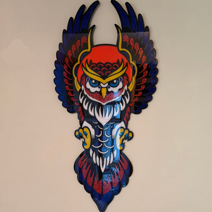 A colorful owl wall art piece mounted on a beige wall, featuring a stylized owl with dark blue wings, a red and yellow head, white feathers, and blue and red body details.