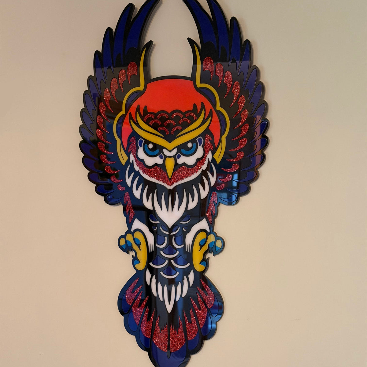 A colorful owl wall art piece mounted on a beige wall, featuring a stylized owl with outstretched wings in red, blue, yellow, and white.