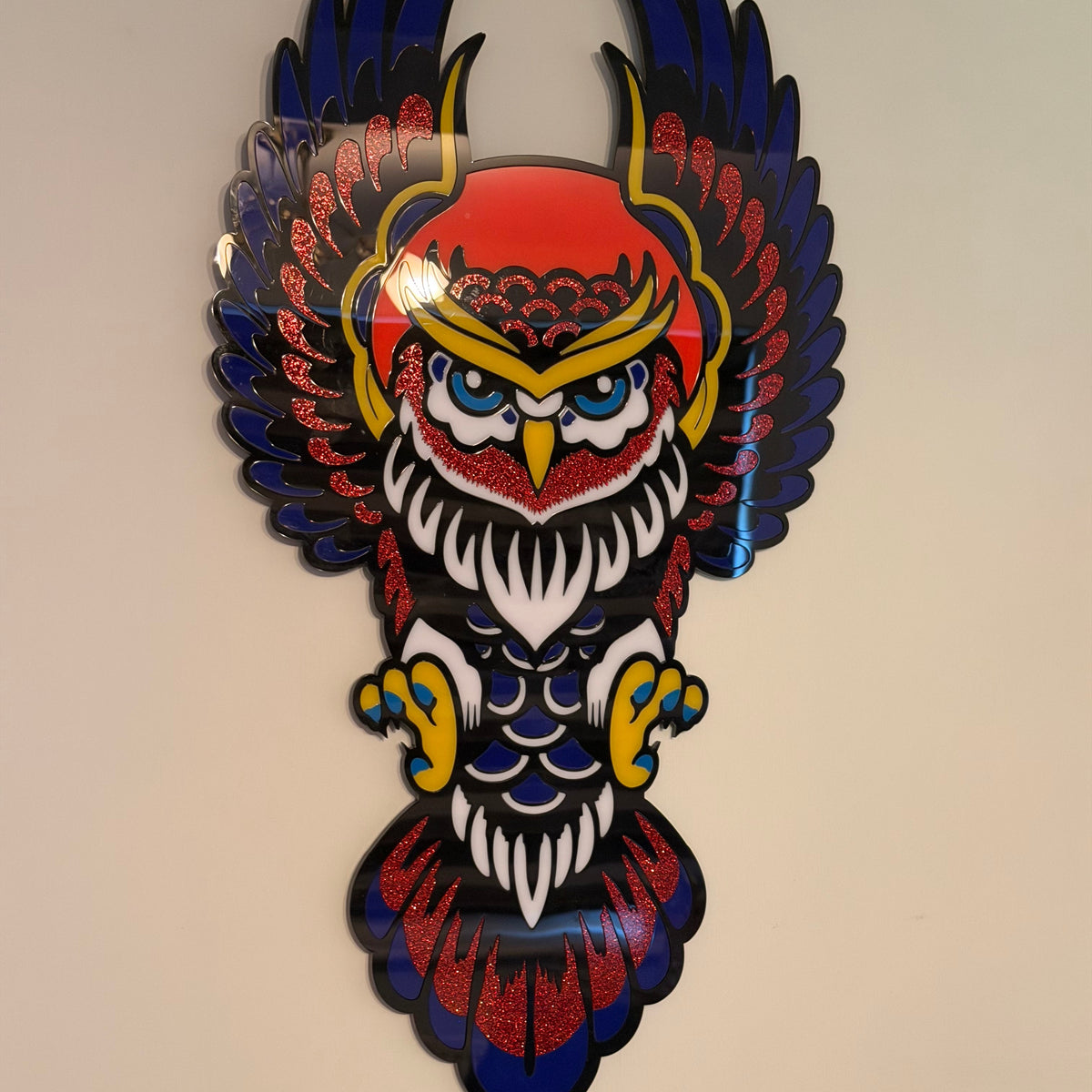 A colorful owl wall art piece with outstretched wings, featuring a red head, blue and black wings, white feathers, and yellow accents, mounted on a beige wall.