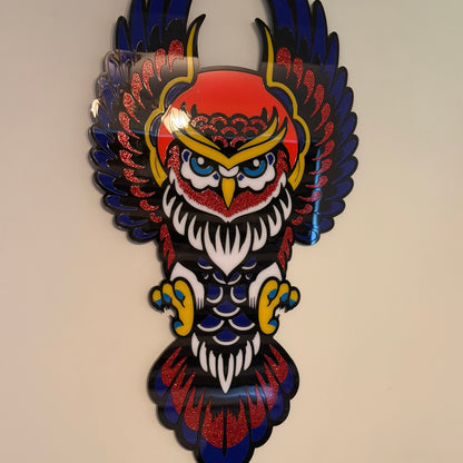 A colorful owl wall art piece with outstretched wings, featuring a red head, blue and black wings, white feathers, and yellow accents, mounted on a beige wall.
